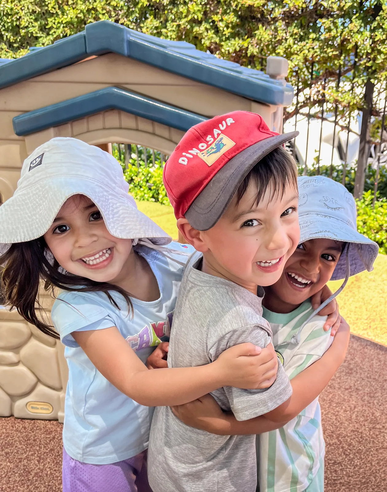 Three Starbright students in sun hats hugging and laughing together outside by the playhouse